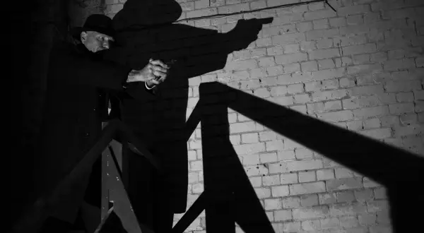 A man in a fedora and trench coat aims a pistol in this dramatic film noir photography. His shadow looms large against a brick wall.