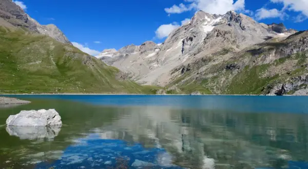 Le lac de la Sassière, aux eaux turquoise, reflète les montagnes majestueuses et le ciel bleu. Un rocher émerge, témoin de la quiétude du paysage.