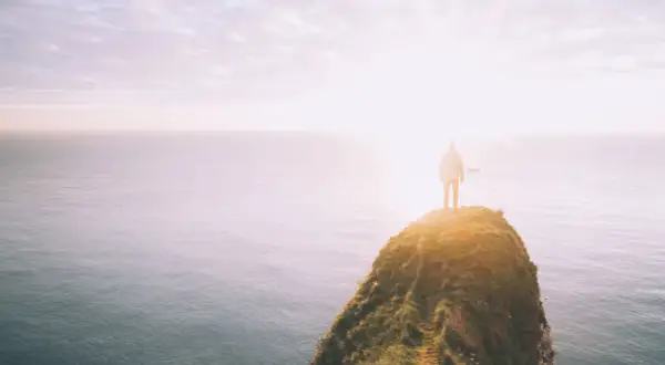 Ein Mensch steht auf einer Klippe am Meer, die Sonne scheint hell. Ein Symbol für das Durchhalten in schweren Zeiten, wenn man dem Horizont entgegenblickt.