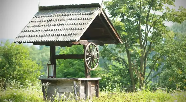 Stara studnia z drewnianym kołem i dachem, otoczona polnymi kwiatami. Zastanawiasz się, ile kosztuje wykopanie studni głębokości 25 m?