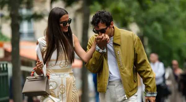 Ein Mann küsst die Hand einer Frau. Sein Outfit für ein erstes Date: gelbe Jacke, weißes T-Shirt, helle Hose. Sie trägt ein gemustertes Kleid und eine Tasche.