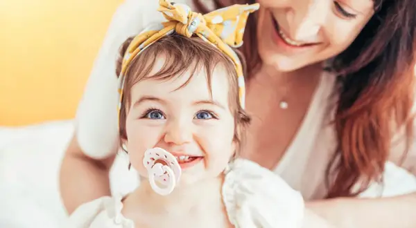 A baby with bright blue eyes and a yellow headband smiles, her passy teeth peeking out from behind a pacifier. Her mother smiles in the background.