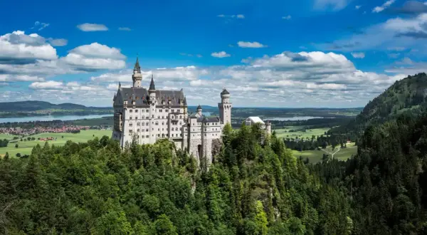 El castillo del rey loco, Neuschwanstein, se alza majestuoso sobre un risco verde, con torres que acarician las nubes y un paisaje de ensueño.