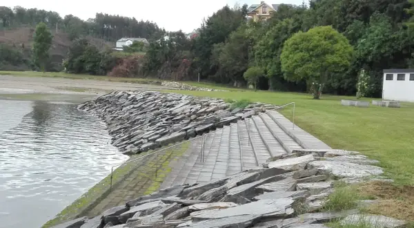 Escaleras de piedra y césped verde junto al agua en la playa de los bloques Ribadeo, con casas y árboles al fondo.