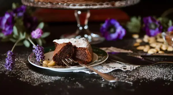 Ein Stück saftiger italienischer Schokokuchen, bestäubt mit Puderzucker, auf einem Teller mit Mandeln und lila Blumen.