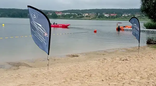 Plaża nad jeziorem, czerwona łódź ratownicza WOPR, pomarańczowa łódź, flagi, budynki na horyzoncie.
