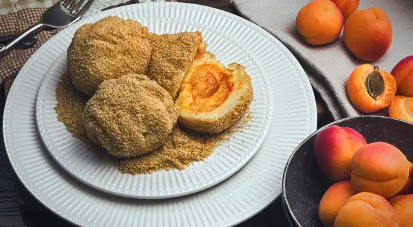 Mehlspeisen aus Kartoffelteig mit Aprikosenfüllung, paniert und auf einem Teller mit zerbröselten Semmelbröseln angerichtet. Daneben frische Aprikosen.