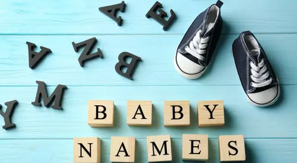 Des lettres noires forment "BABY NAMES" sur des cubes en bois. Une paire de chaussons bleus attend. Quel joli prénom en M pour le bébé ?