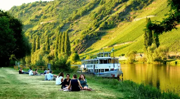 Entspannte Menschen genießen die Sonne am Ufer der Mosel. Ein Ausflugsschiff liegt vor, im Hintergrund steile Weinberge. Perfekte Mosel Ausflugsziele.