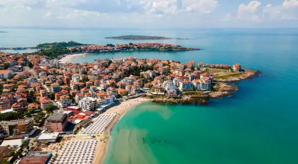 Malownicze wybrzeże Bułgarii nad turkusowym morzem. Miasto z czerwonymi dachami rozciąga się wzdłuż piaszczystej plaży z białymi parasolami.