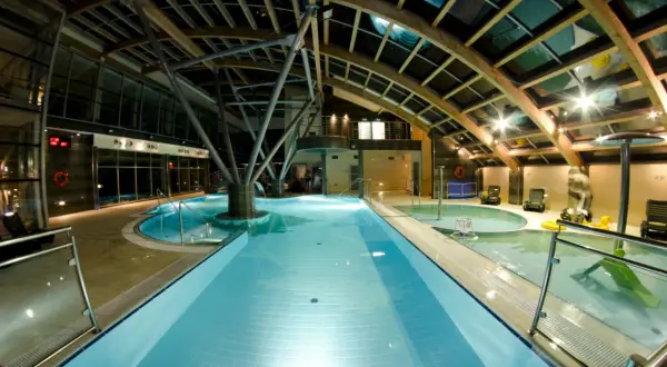Wnętrze Laguna Kompleks Wypoczynkowo Rekreacyjny Mrzeżyno: baseny, jacuzzi, leżaki pod drewnianą, przeszkloną konstrukcją dachu.