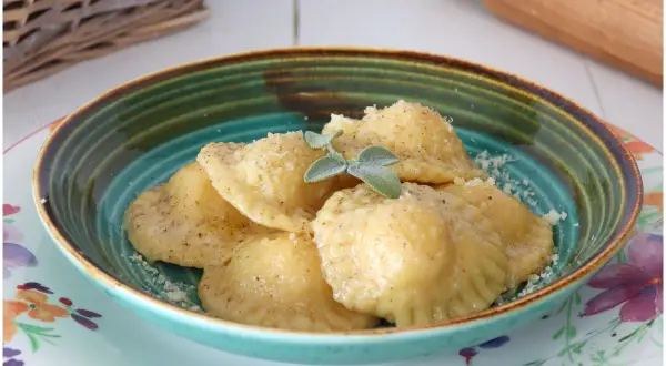 Tortelli di patate fumanti, cosparsi di parmigiano e guarniti con salvia, in una ciotola colorata.