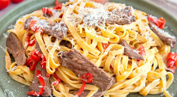 Herzhaftes Gericht mit Pasta und zarten Rinderfiletstreifen, garniert mit geriebenem Parmesan und roten Paprikastücken.