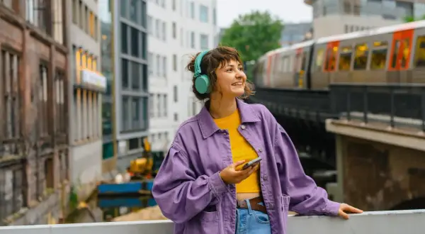 Junge Frau mit Kopfhörern und Handy in der Hand, die auf eine Stadtkulisse mit Zug blickt. Sie plant ihr Fachhochschulreife Fernstudium.