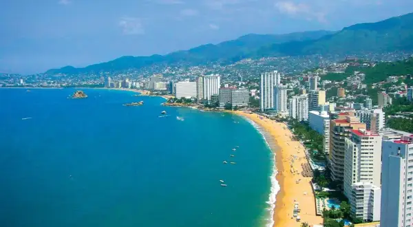 Słoneczna plaża w Acapulco z turkusową wodą, białymi hotelami i zielonymi wzgórzami w tle.