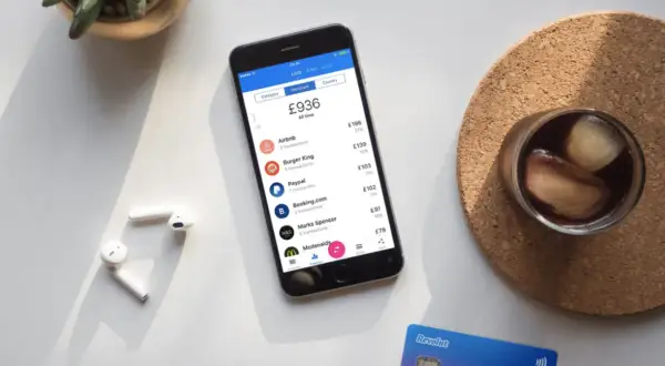 Telefon z aplikacją Revolut, pokazujący wydatki i opcje, np. revolut ubezpieczenie podróżne. Obok słuchawki i drink.