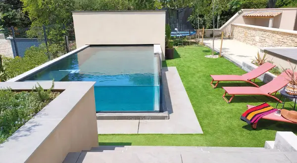 Un magnifique bassin vitré aux eaux bleues, entouré d'une pelouse synthétique et de chaises longues roses.