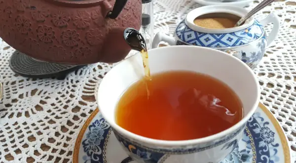 Ciemnobrązowy kolor herbaty w filiżance, nalewanej z ozdobnego czajniczka. Obok cukiernica z brązowym cukrem.