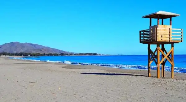 Torre de vigilancia en la vera playa, con el mar azul y montañas al fondo.