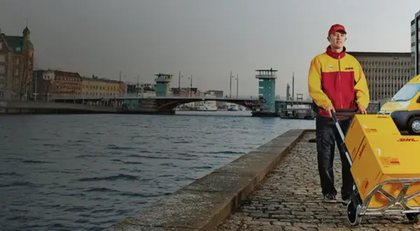 Kurier DHL z wózkiem i paczką do Włoch na tle kanału i miasta.
