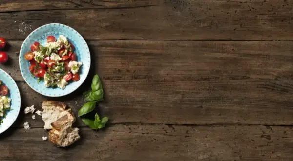 Två tallrikar med tomatsallad och vispad mozzarella, garnerad med basilika. Körsbärstomater och bröd på träbord.