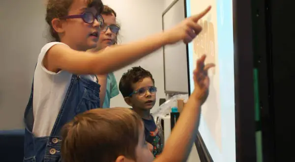 Kinder mit Brillen lernen interaktiv an einem Touchscreen. Sie zeigen auf Zahlen und Symbole, was multisensorisches Lernen ermöglicht.