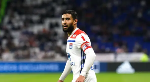 Piłkarz Nabil Fekir w białej koszulce z czerwonymi elementami, z opaską kapitańską na ramieniu, na tle trybun.