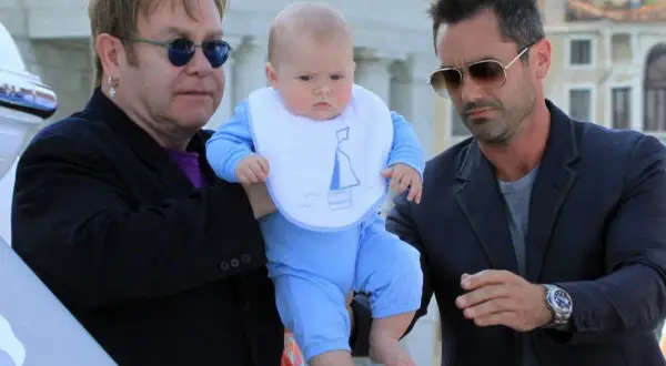 Elton John z dziećmi, w tym małym dzieckiem w niebieskim ubranku i śliniaczku z łódką, w otoczeniu innych mężczyzn.
