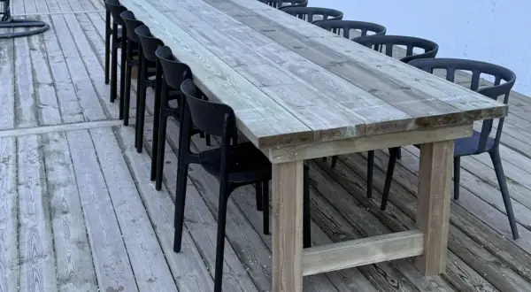 Ett stadigt, hemmabyggt utebord av trä med plats för många gäster. Svarta stolar står uppradade runt det robusta bordet på altanen.