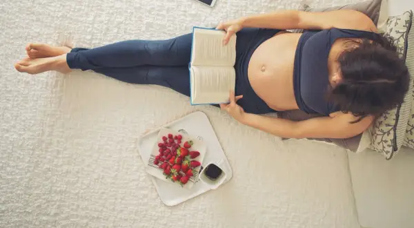 Ciąża 22 tydzień to już piąty miesiąc. Kobieta leży na łóżku, czyta książkę, obok taca z owocami i kawą.