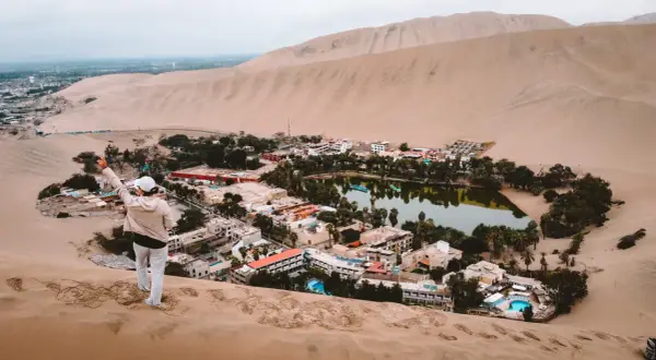 Oaza na pustyni Huacachina, z turystką na wydmie pozdrawiającą zieloną, bujną okolicę z jeziorem i hotelami.
