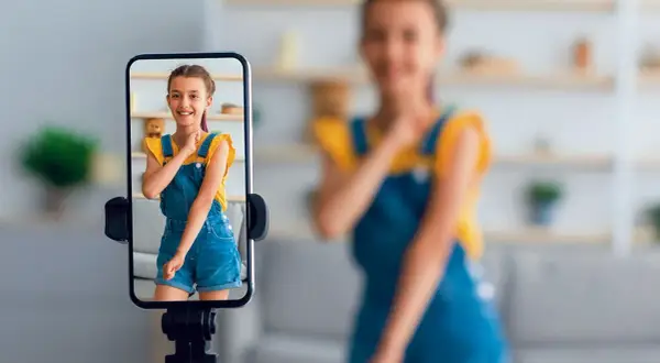 Dziewczynka na ekranie telefonu uśmiecha się, podczas gdy mama w tle nagrywa filmik. To przykład zjawiska sharenting co to znaczy dzielenie się życiem dzieci w sieci.