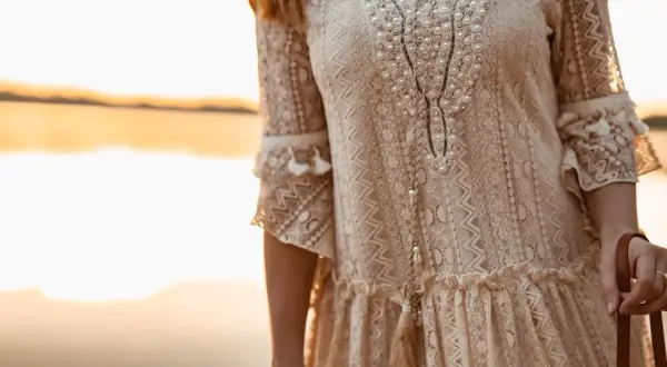 Kobieta w koronkowej sukience w stylu boho, ozdobionej perłami i frędzlami, na tle zachodzącego słońca.