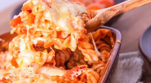 Un cucchiaio solleva una porzione generosa di pasta alla sorrentina, filante e dorata, da una pirofila marrone.