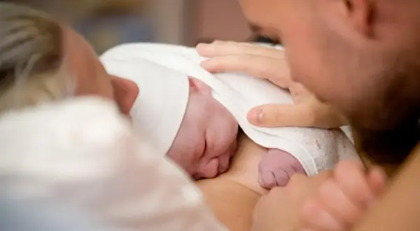 Pierwsze chwile po narodzinach. Noworodek otulony kocykiem, rodzice z czułością dotykają maleńkiego ciałka. Tak wygląda poród.