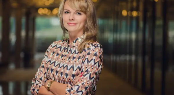 Portret uśmiechniętej kobiety w kolorowej bluzce, z rękami skrzyżowanymi na piersi.