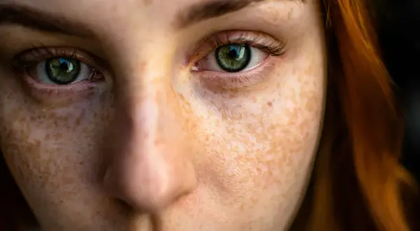 Close-up texture photography of a young woman's face, highlighting her green eyes and the intricate pattern of her freckles.