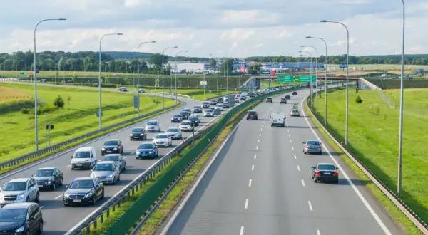 Autostrada z ruchem samochodowym w słoneczny dzień, z zielonymi poboczami i drzewami w tle.