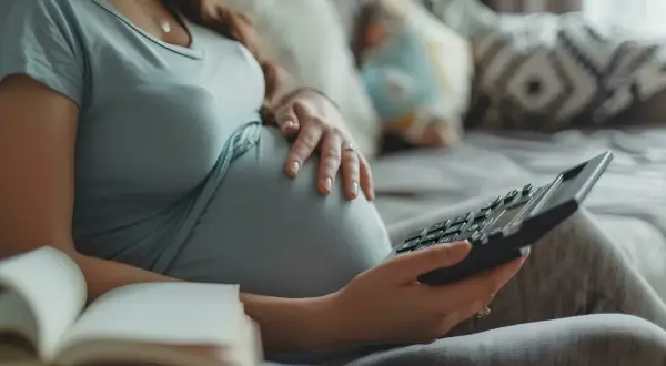 Ciężarna kobieta z kalkulatorem i otwartą książką na kanapie.