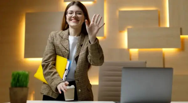 Uśmiechnięta kobieta w okularach, z teczkami i kawą, macha ręką. To jej dzień próbny w pracy bez umowy, pełen nadziei.