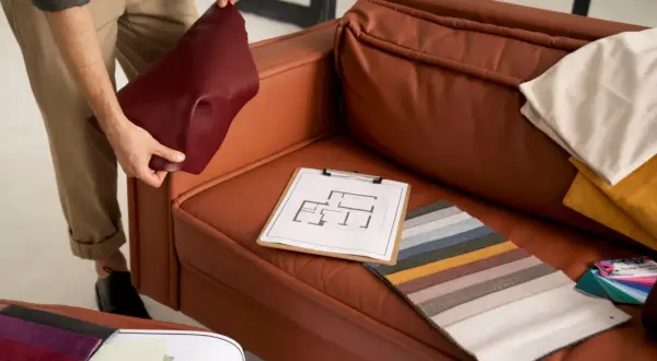 A person is choosing couch materials, holding a burgundy swatch next to a brown leather couch. Fabric samples and a floor plan are on the seat.