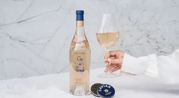 Une main tient un verre de vin rosé. À côté, une bouteille de Clos du Temple et une boîte de caviar. Une belle conservation rosé pour un moment d'exception.