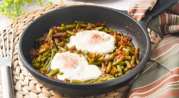 Judías verdes salteadas con trocitos de bacon y dos huevos fritos encima, servido en sartén.