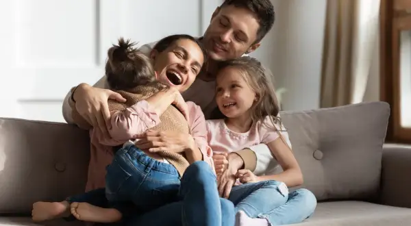Szczęśliwa rodzina na kanapie. Mama śmieje się, tuląc córkę. Tata obejmuje drugą córkę. To piękny obraz rodziny, gdzie każdy czuje się kochany, nawet jeśli ktoś jest pasierbem, kto to nie ma znaczenia w tej miłości.