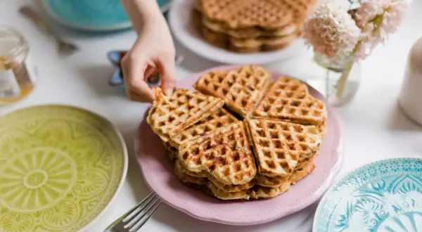 Ein Kind greift nach einem herzförmigen Waffel ohne Ei. Mehrere Stapel Waffeln liegen auf rosa und weißen Tellern, umgeben von bunten Tellern und Blumen.