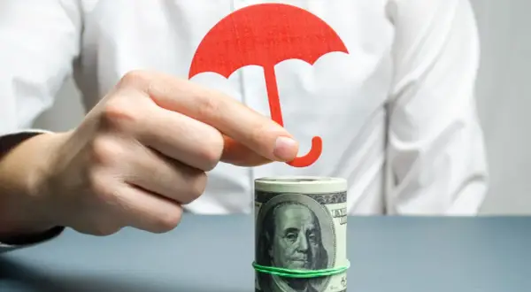 Dłoń trzyma czerwony parasol nad zrolowanymi dolarami. Tani murek oporowy dla Twoich finansów.
