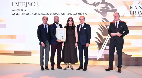 Pięć osób w garniturach i sukience stoi na scenie, odbierając nagrodę.