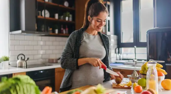 Uśmiechnięta ciężarna kobieta przygotowuje zdrowy posiłek, dbając o dietę w ciąży. Na blacie świeże warzywa i owoce.