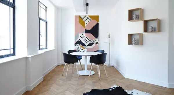 Un comedor moderno con mesa redonda, sillas negras y una alfombra geométrica. Las estanterías flotantes y las ventanas grandes dan la sensación de casas pequeñas y acogedoras.