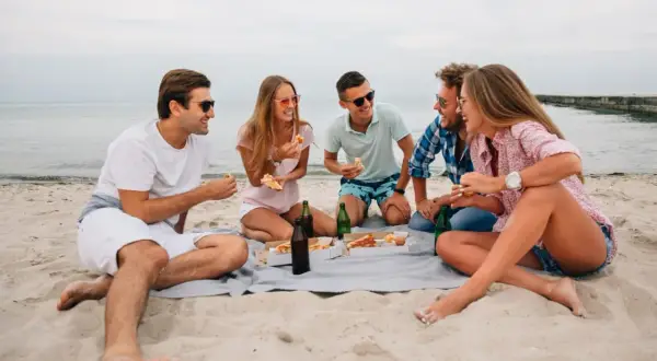 Grupa przyjaciół cieszy się piknikiem na plaży, jedząc pizzę i pijąc napoje. To świetny pomysł na wakacje!